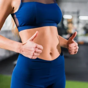 Femme en haut de sport bleu faisant un pouce levé, exprimant la satisfaction après un entraînement pour perdre du gras abdominal.