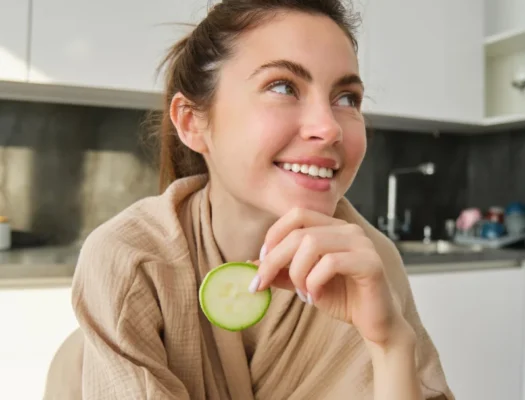 A woman with dull skin smiles while holding a cucumber in her hand, showcasing a cheerful expression.