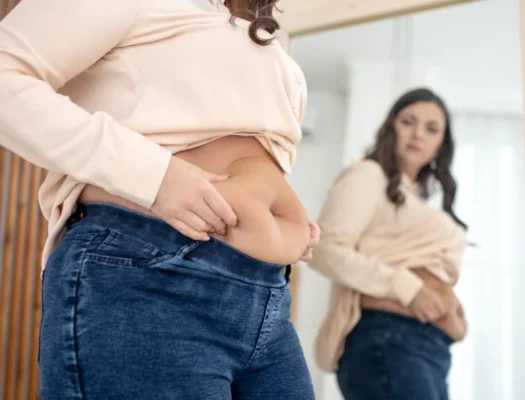 A woman gazes at her stomach in the mirror, observing her belly cellulite with a contemplative expression.