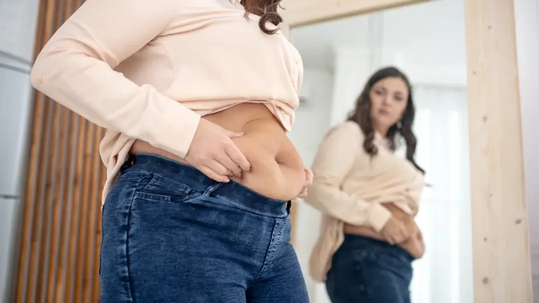Une femme observe son ventre dans le miroir, mettant en évidence la cellulite sur sa peau.